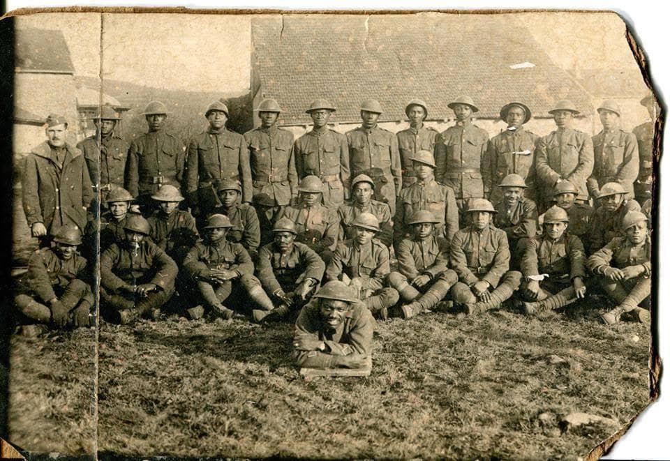 Aged black-and-white photograph of roughly twenty-five soldiers posed in two rows in front of a building with a sloped roof; one man reclines on the ground in front.