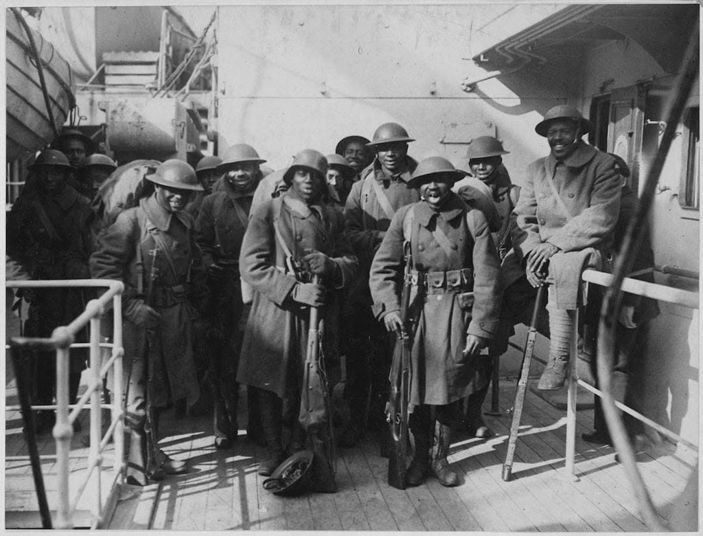 Black-and-white photograph of about fifteen soldiers in overcoats and helmets, some carrying rifles, smiling on the deck of a ship, with railings and lifeboat davits visible.