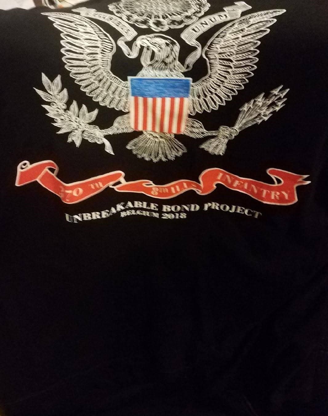 Black T-shirt printed with the Great Seal of the United States eagle emblem and a red banner reading "370TH" and "INFANTRY," with "UNBREAKABLE BOND PROJECT, BELGIUM 2018" printed below.
