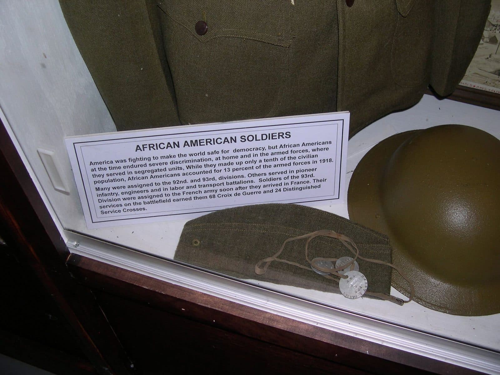Museum display case showing a garrison cap with dog tags on a chain and a steel helmet, beside the full "African American Soldiers" placard text and part of the mounted uniform jacket.
