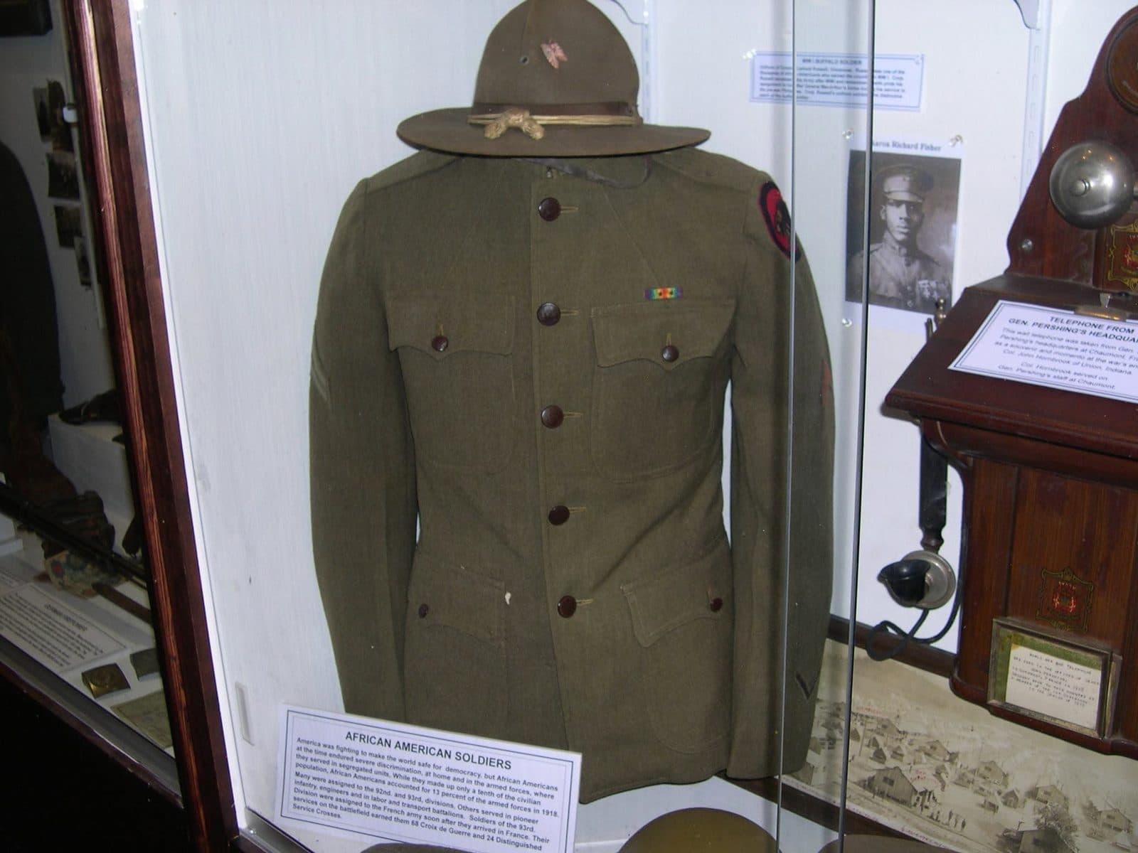 Museum display case showing a World War I-era uniform jacket and campaign hat mounted on a form, beside a photograph and an antique wall telephone, with an "African American Soldiers" placard.