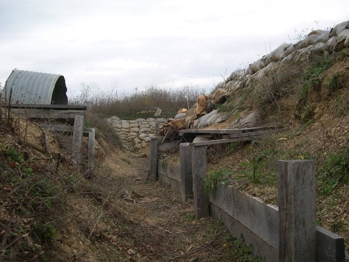 Modern color photograph of an empty preserved trench and dugout site, with a corrugated-metal shelter, sandbagged walls, and wooden revetments, no people present.