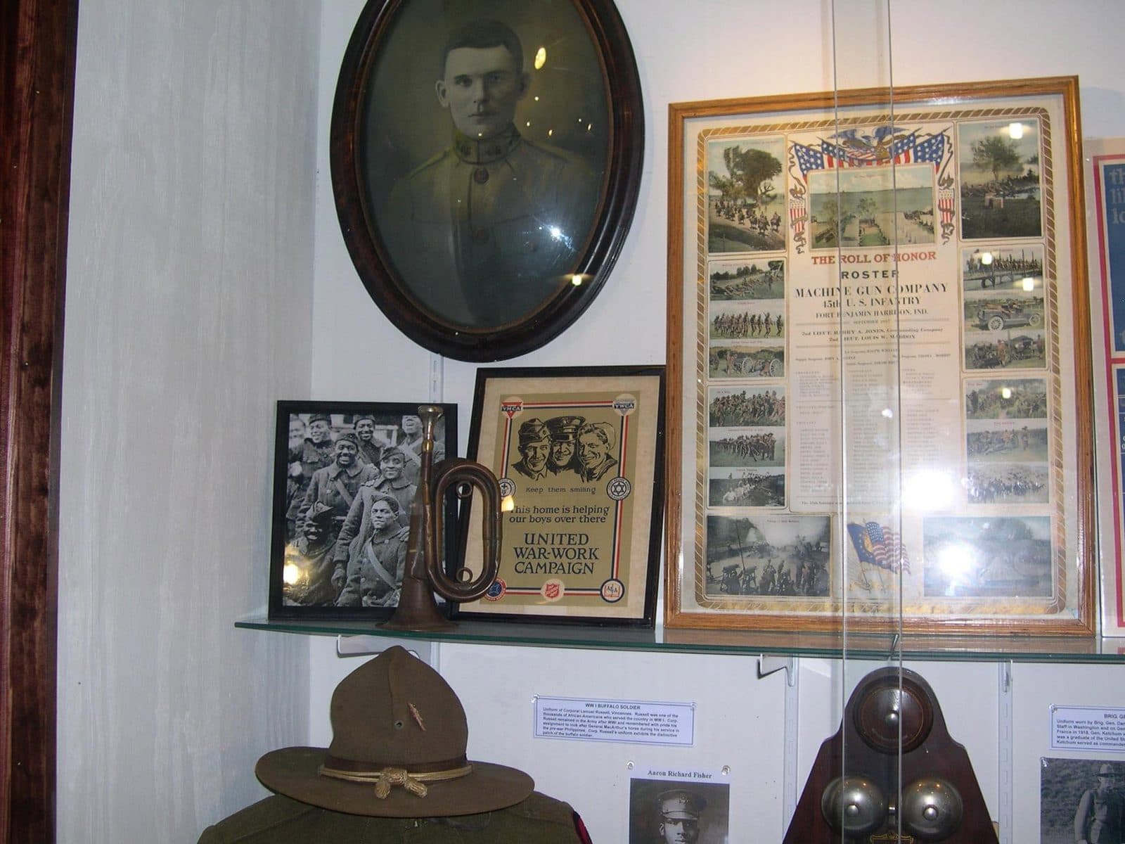 Museum wall display with an oval-framed portrait, a framed roster document, a small group photograph, a bugle, and a poster, arranged above a shelf.
