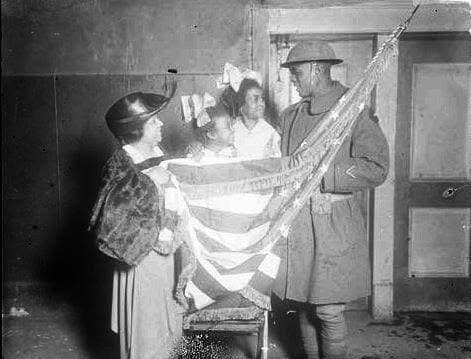 Black-and-white photograph of a soldier and two women holding a large folded American flag between them, in a plain room with a doorway.