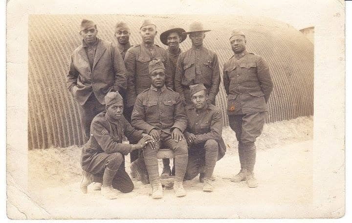 Aged photograph of eight soldiers posed in front of a curved corrugated-metal hut, five standing and three kneeling in front.