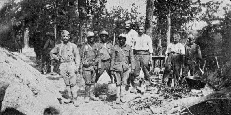 Aged photograph of nine men in a wooded clearing, some in military uniform and some in civilian shirtsleeves, near buckets and a cooking pot.