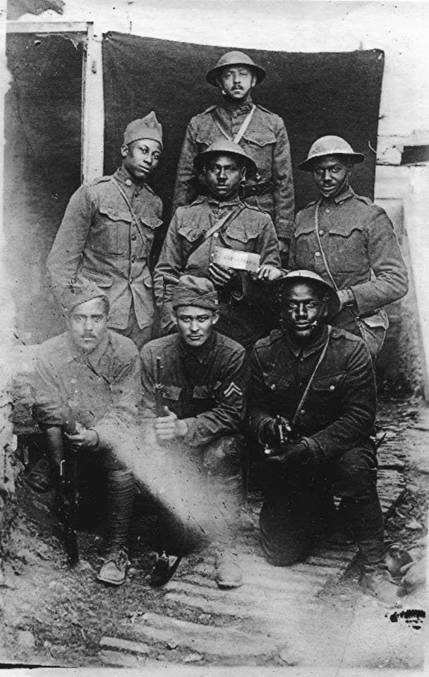 Black-and-white World War I photograph of seven soldiers in helmets and overcoats posed in front of a dark canvas backdrop, on bare ground.