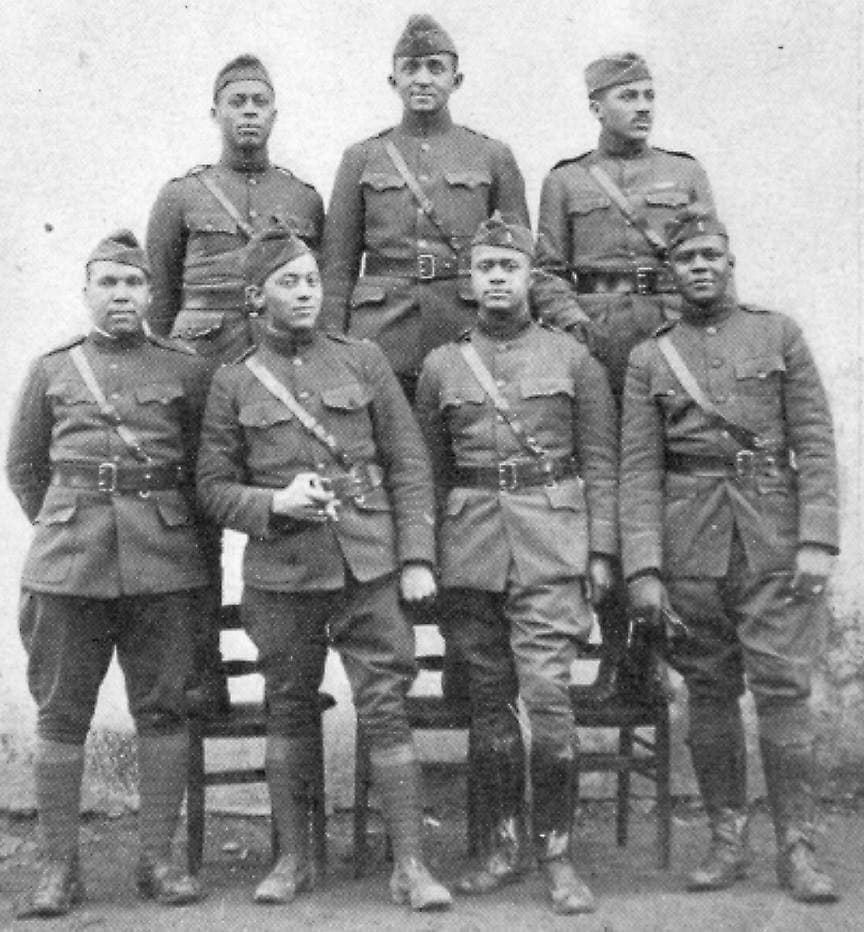 Black-and-white photograph of seven soldiers, four standing and three seated on chairs, posed against a plain wall.