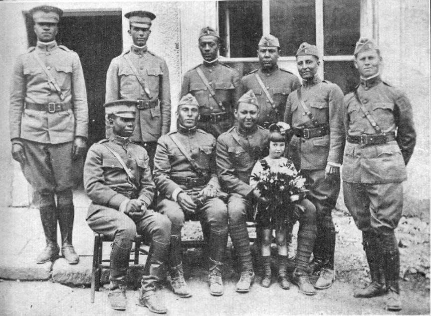 Black-and-white photograph of eight soldiers posed outside a building doorway with a young girl in front holding a bouquet of flowers.