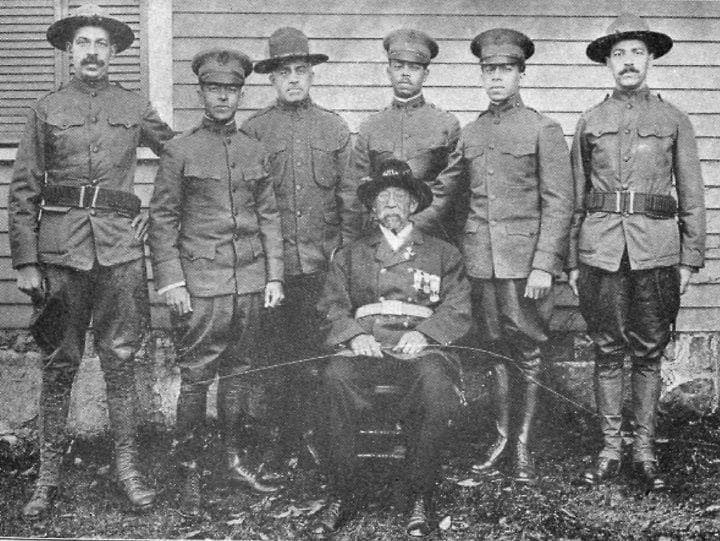 Black-and-white photograph of six soldiers standing behind an older man in a wide-brimmed hat, seated in a chair and wearing service ribbons, in front of a house.