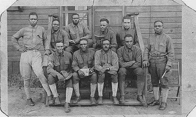 Black-and-white photograph of ten soldiers, some standing and some seated on wooden steps, in front of a wooden building.
