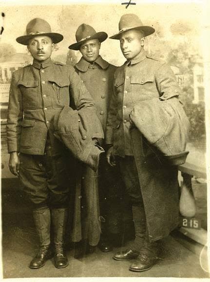 Studio portrait of three soldiers in campaign hats and overcoats posed in front of a painted scenic backdrop.