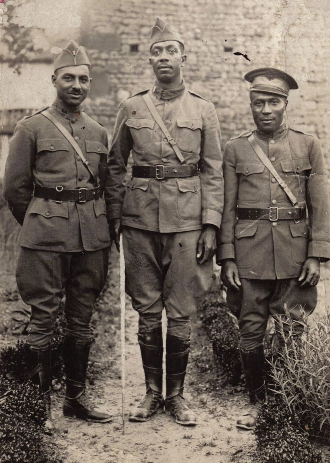 Full-length photograph of three soldiers standing in front of a stone wall, one leaning on a cane.