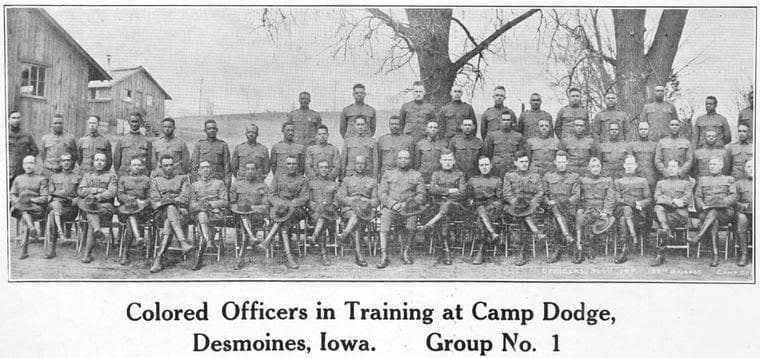Black-and-white photograph of a large group of soldiers in two rows, seated and standing, in front of wooden buildings and a bare tree.