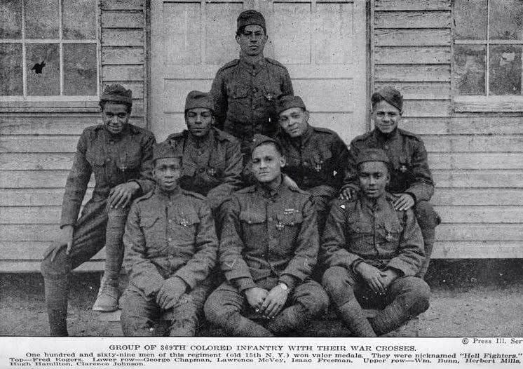 Black-and-white photograph of eight soldiers posed in front of a wooden building, four standing and four seated, wearing medals.