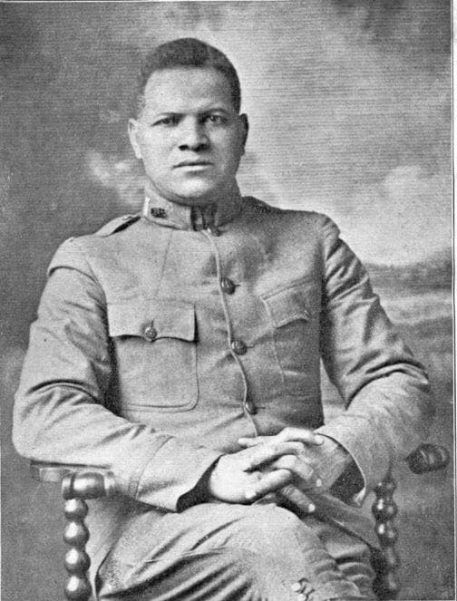 Studio portrait of a soldier seated in a chair, hands clasped, wearing a service coat, against a painted studio backdrop.