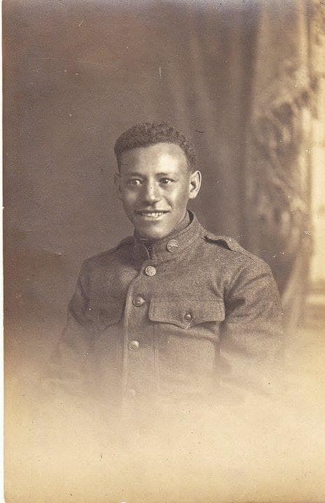 Studio portrait of a young soldier in uniform, smiling, against a draped curtain backdrop.