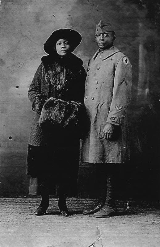 Studio portrait of a soldier in an overcoat standing beside a woman in a fur-trimmed coat and wide-brimmed hat, holding a fur muff.