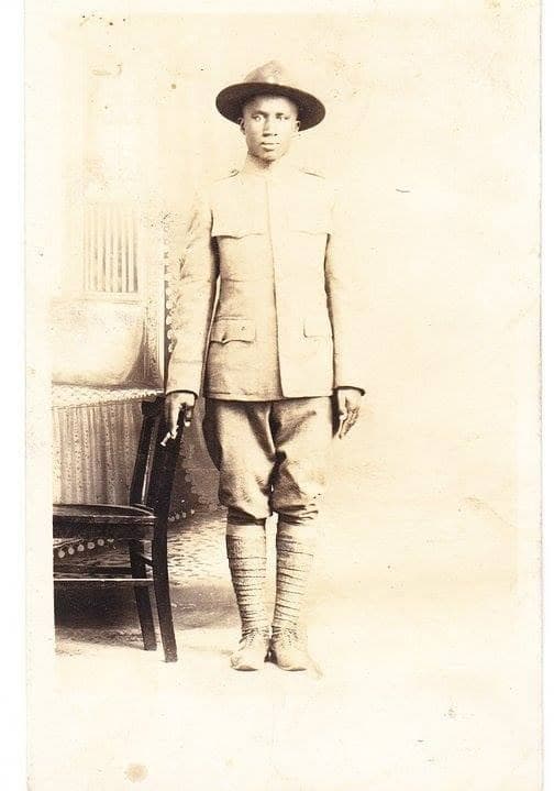 Studio portrait of a soldier in a garrison cap and full uniform with puttees, standing beside a wooden chair.