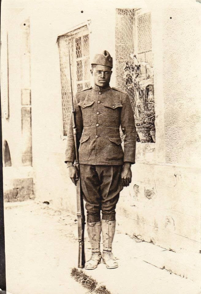 Full-length photograph of a soldier standing at attention with a rifle beside a stucco building with wire-mesh window screens.