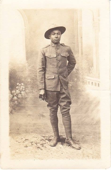 Studio portrait of a young soldier in a campaign hat and full uniform, standing in front of a painted garden backdrop with an arch.
