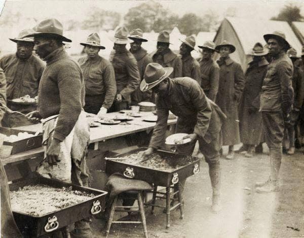 Black-and-white photograph of soldiers in campaign hats at outdoor serving tables with food trays, with rows of tents visible behind them.