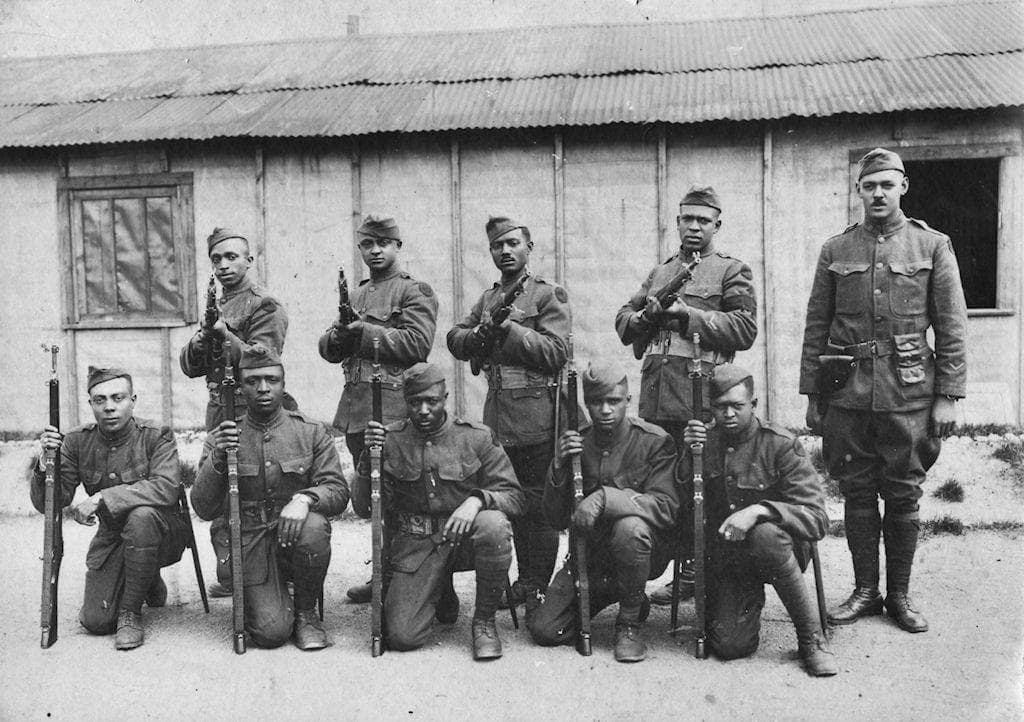 Black-and-white photograph of nine soldiers holding rifles with fixed bayonets, kneeling and standing in two rows in front of a corrugated barracks building, with an officer standing apart at right.