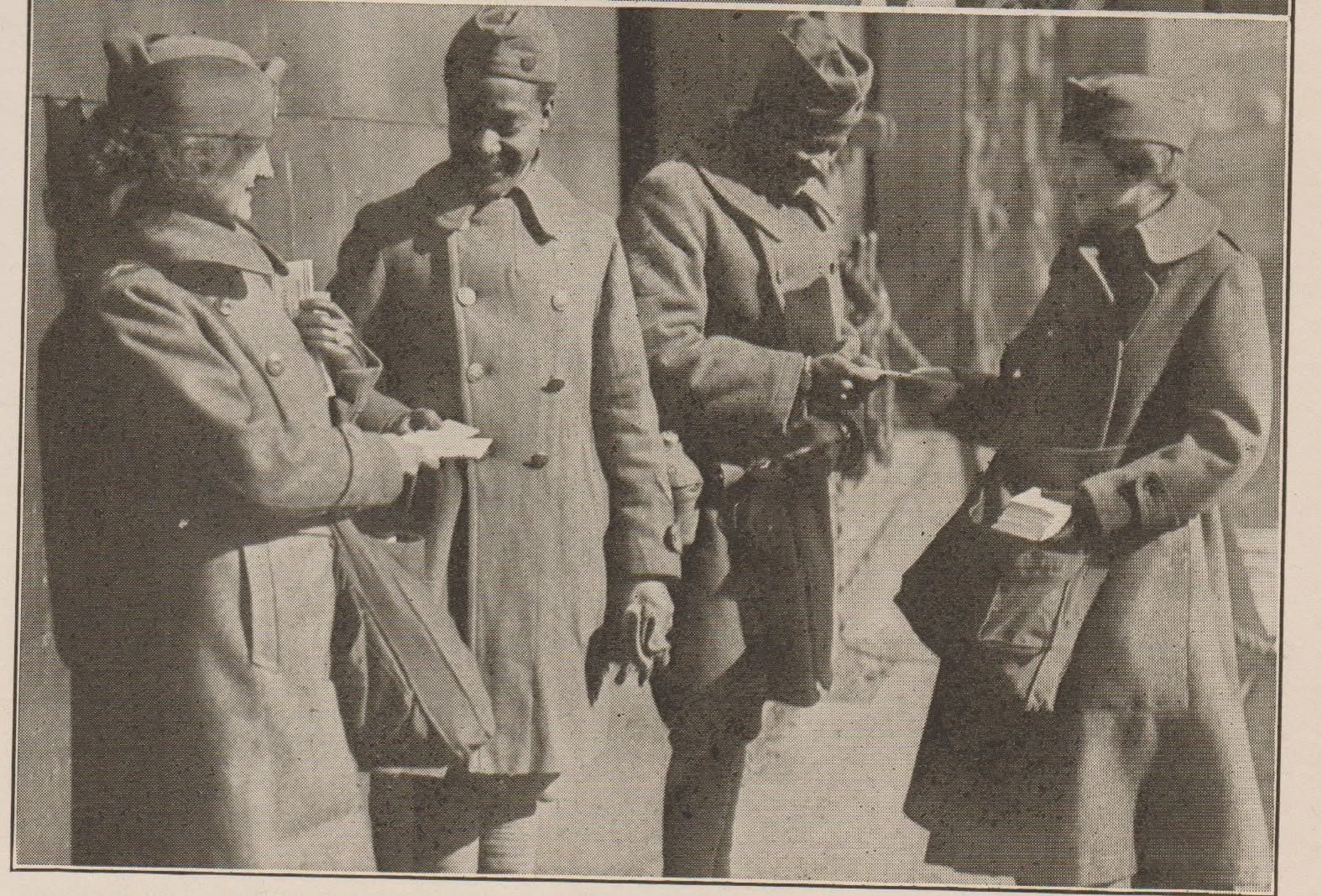Aged publication photograph of four people in long coats and garrison caps on a street, exchanging papers by hand.