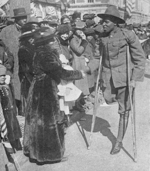 Black-and-white photograph of a soldier on crutches, one leg bandaged, greeting a crowd of women on a city street lined with American flags.