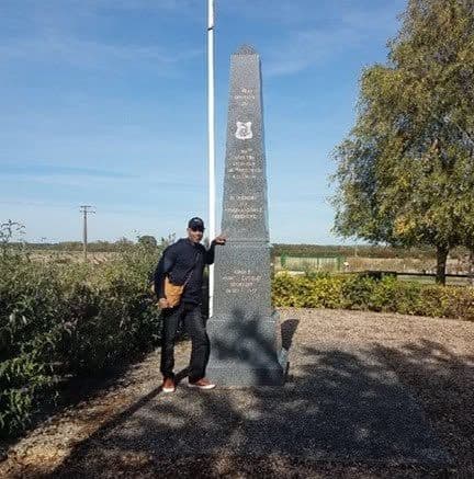 Modern color photograph of a man in dark casual clothing standing beside a gray granite obelisk monument and flagpole, on a paved path near hedges.