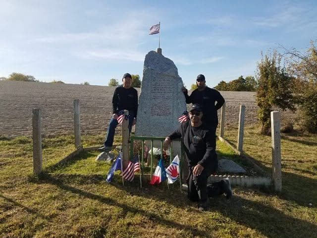 Modern color photograph of three people posed at a stone memorial monument in an open field, American and French flags displayed at its base.