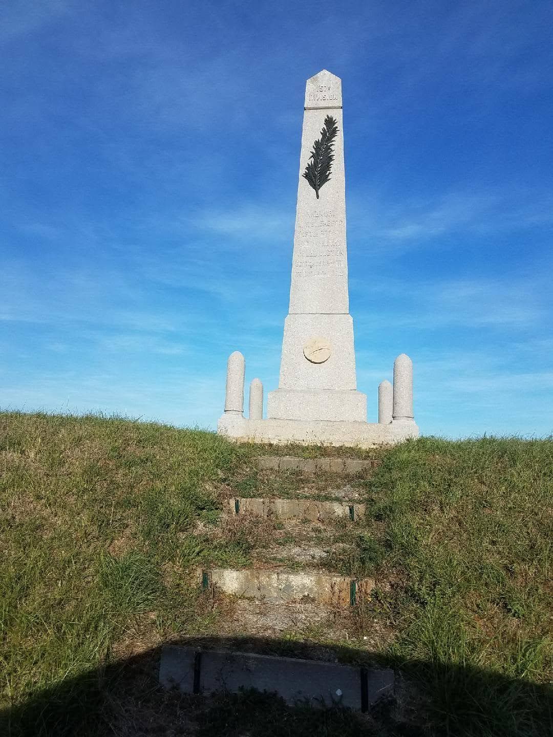 Modern color photograph of a tall stone obelisk monument on a grassy hill under a blue sky, with stone steps leading up to it.