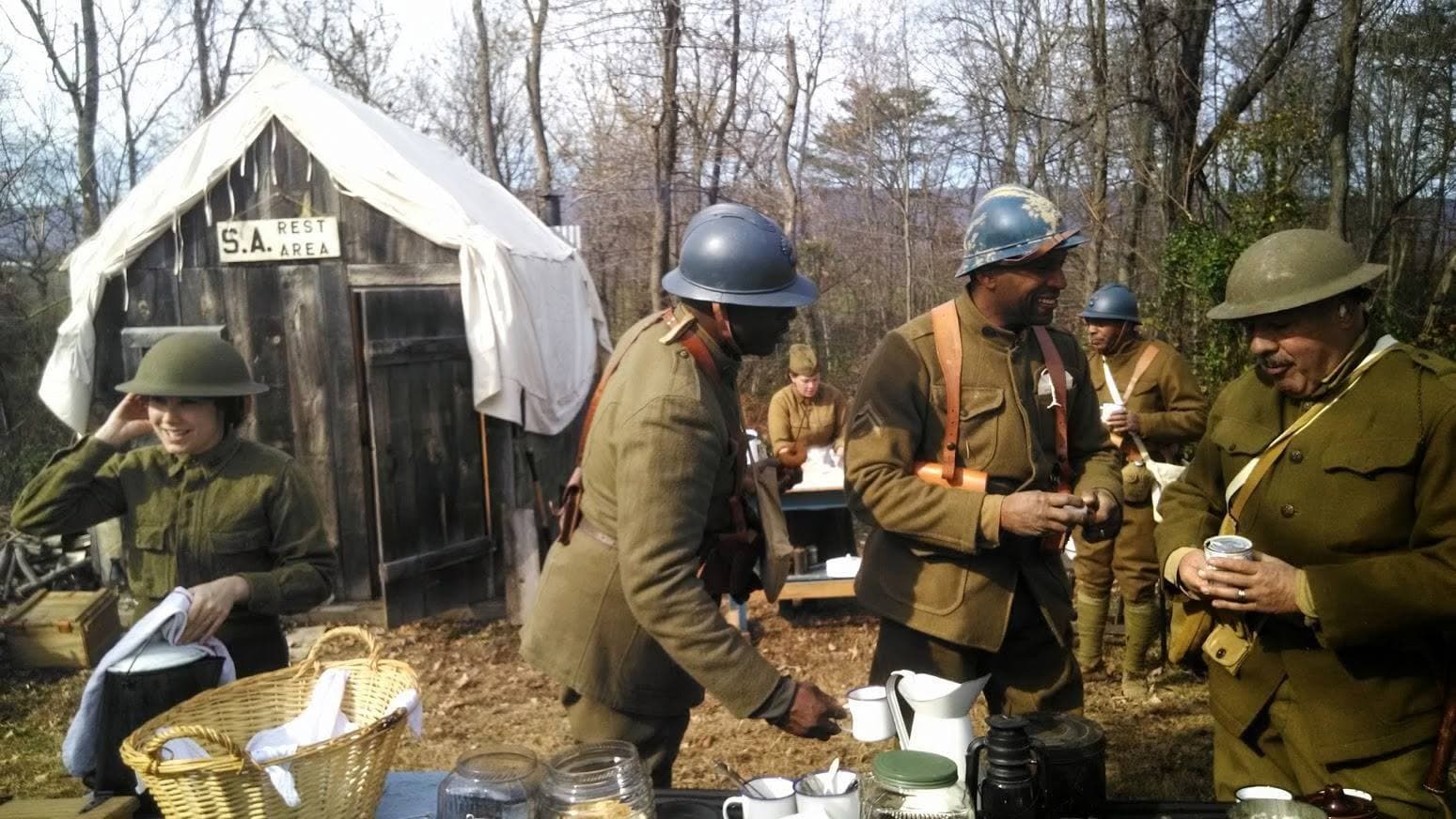 Modern color photograph of reenactors gathered at a rustic wooden shelter with a hand-painted "S.A. Rest Area" sign, some holding drinks near jars and a coffee urn.
