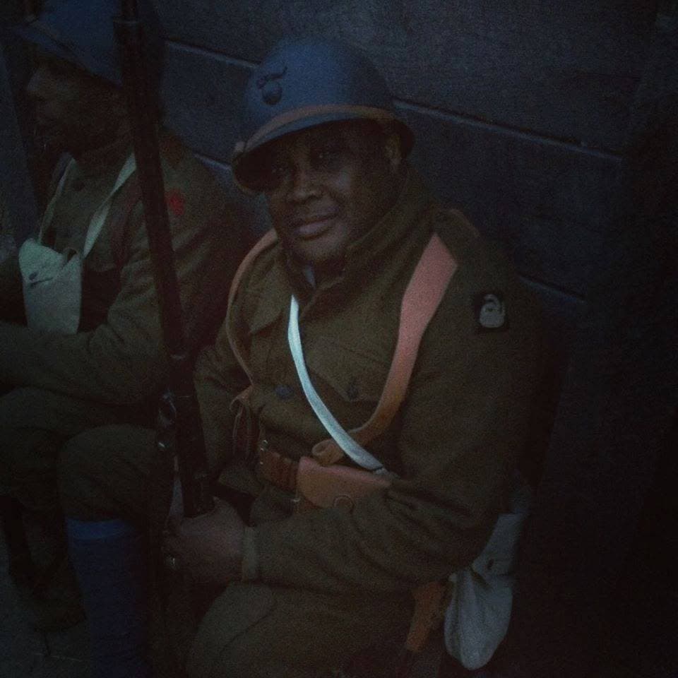 Dimly lit modern color photograph, close portrait of a smiling reenactor seated against a stone wall, another reenactor partly visible in the foreground.