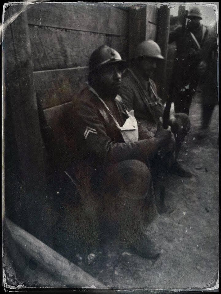 Filtered modern photograph, aged-tone effect, of two reenactors seated against a wood-plank trench wall, one with an arm sling bandage.