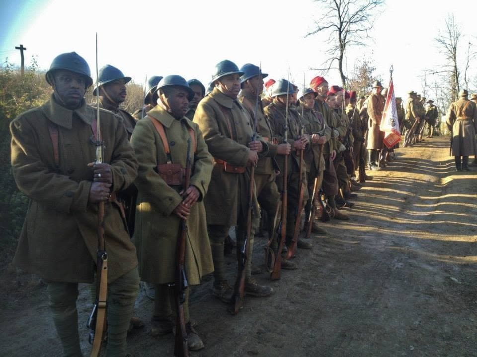 Modern color photograph of a long line of reenactors standing at attention with fixed bayonets along a dirt road, with a color guard flag visible in the distance.
