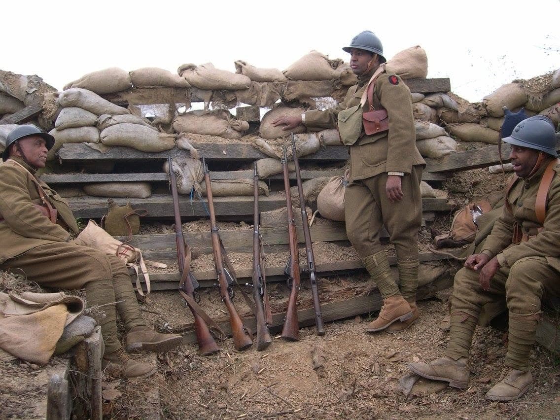 Modern color photograph of three reenactors gathered around a row of rifles stood on end at a sandbagged trench position.