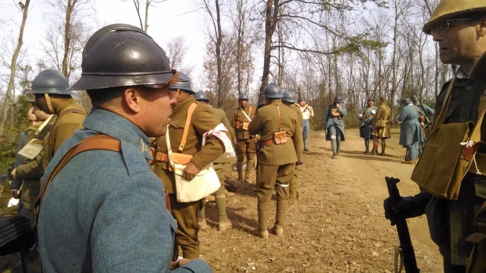 Modern color photograph of reenactors in mixed uniforms, including a blue French-style overcoat, gathered on a wooded dirt path.