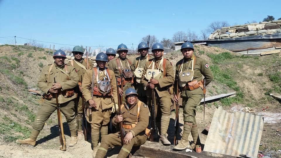 Modern color photograph of nine reenactors posed together at a trench and dugout site with a corrugated-metal shelter, gas mask bags and rifles visible.