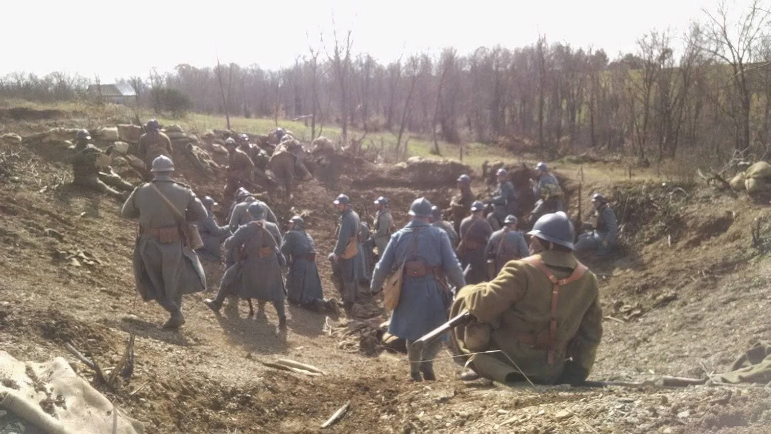 Modern color photograph of a large group of reenactors, several in French horizon-blue overcoats, gathered in a shallow trench and crater area with sandbagged parapets.