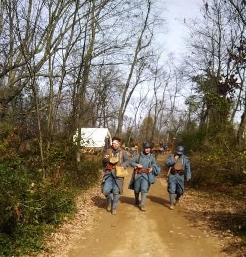 Modern color photograph of three reenactors in French Adrian helmets marching down a wooded dirt path, with a canvas tent visible behind them.