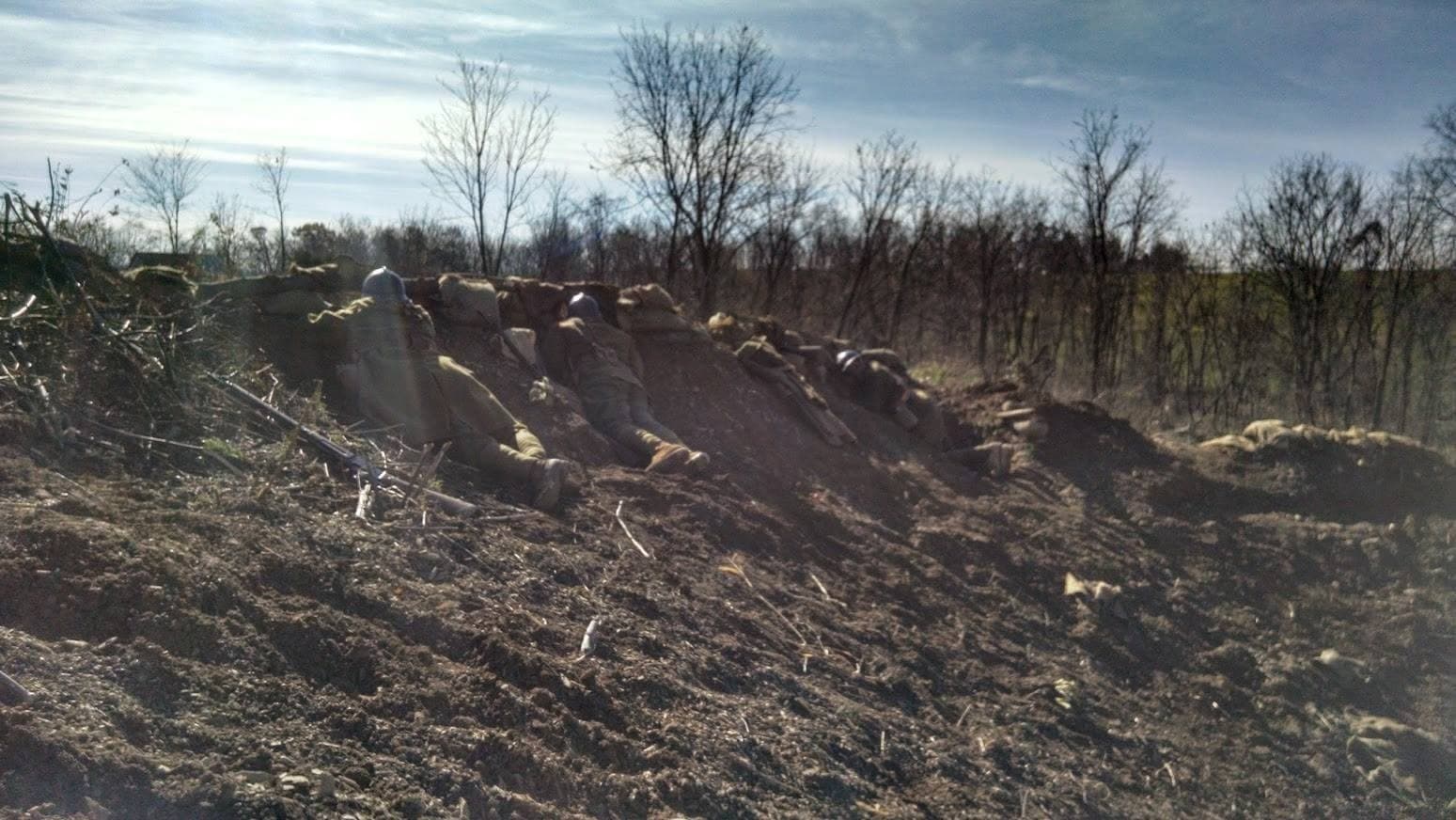 Modern color photograph of reenactors lying prone along a dirt trench embankment with sandbags scattered nearby, bare autumn trees in the background.