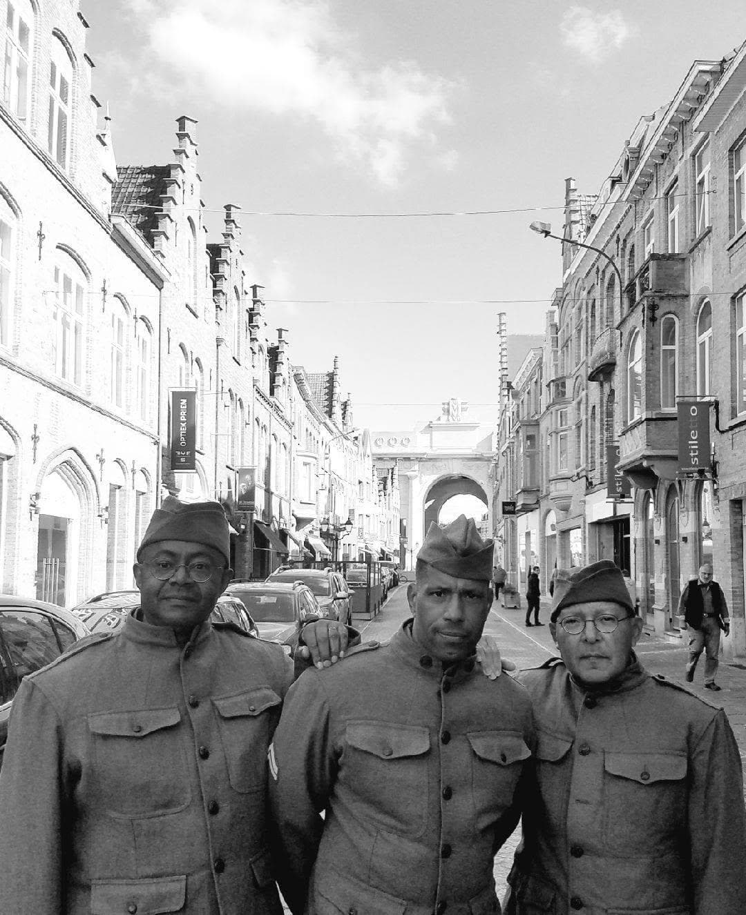 Modern black-and-white photograph of three reenactors in World War I-pattern uniforms on a European street, with a stone archway and rowhouses in the background.