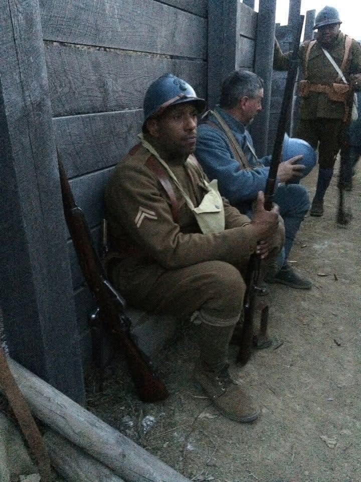 Modern color photograph of reenactors seated against a wooden trench wall, rifles with fixed bayonets propped between their knees, gas mask bags on their chests.