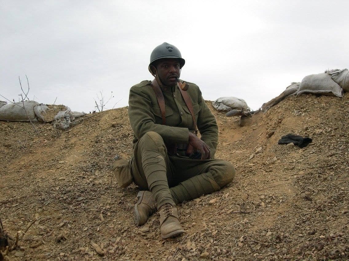 Modern color photograph of a reenactor sitting on the ground in a trench, smoking a pipe, sandbags visible around him.
