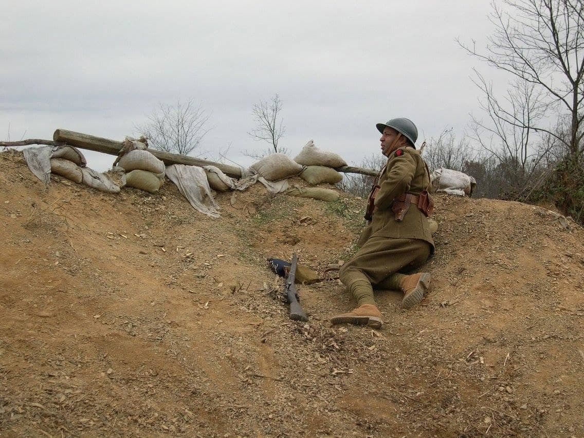 Modern color photograph of a reenactor seated in a trench beside a sandbagged wall, a single rifle propped nearby.