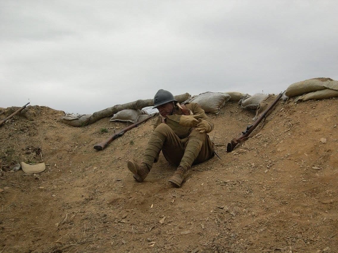 Modern color photograph of a reenactor sliding down a dirt slope near a sandbagged wall, in a dramatic action pose with rifles dropped in the foreground.