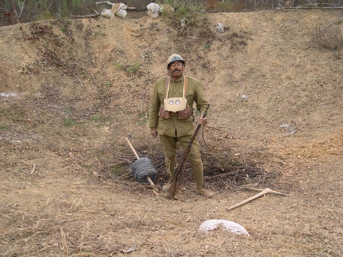 Modern color photograph of a reenactor standing in a shell crater beside a coil of barbed wire and wooden wire pickets, rifle in hand.