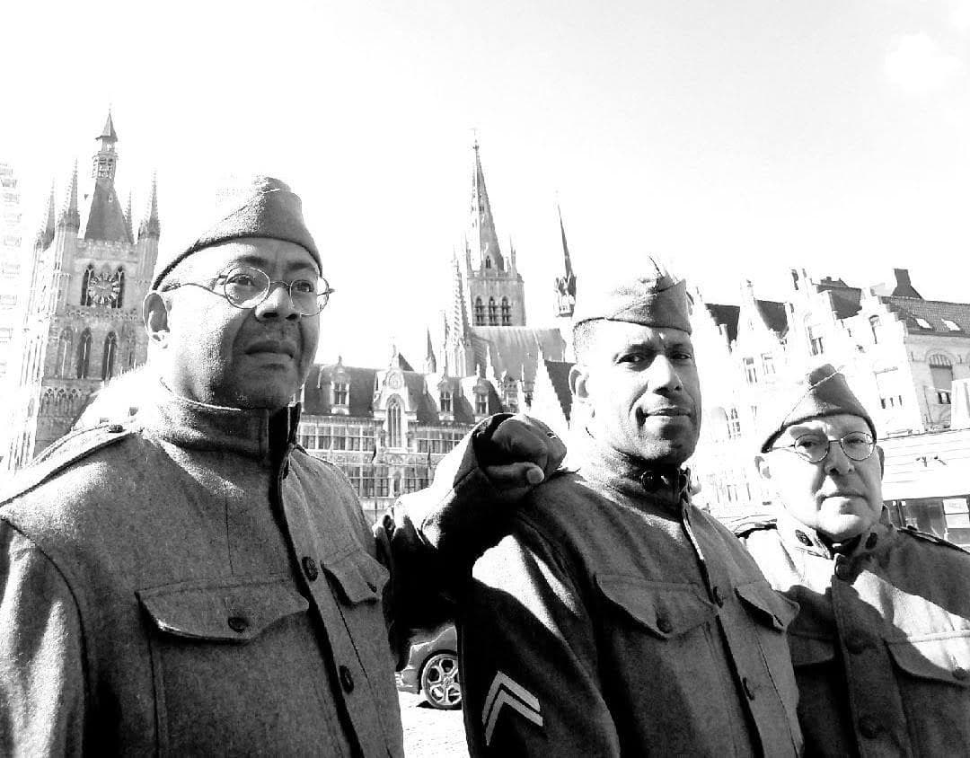 Modern black-and-white photograph of three reenactors in World War I-pattern uniforms and garrison caps standing in a European town square with Gothic church spires in the background.