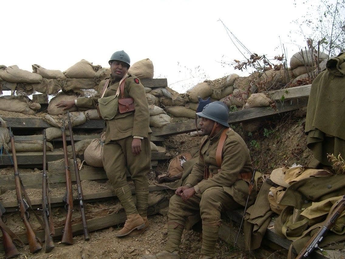 Modern color photograph of two reenactors, one standing and one seated, talking in a trench lined with stacked rifles and sandbags.